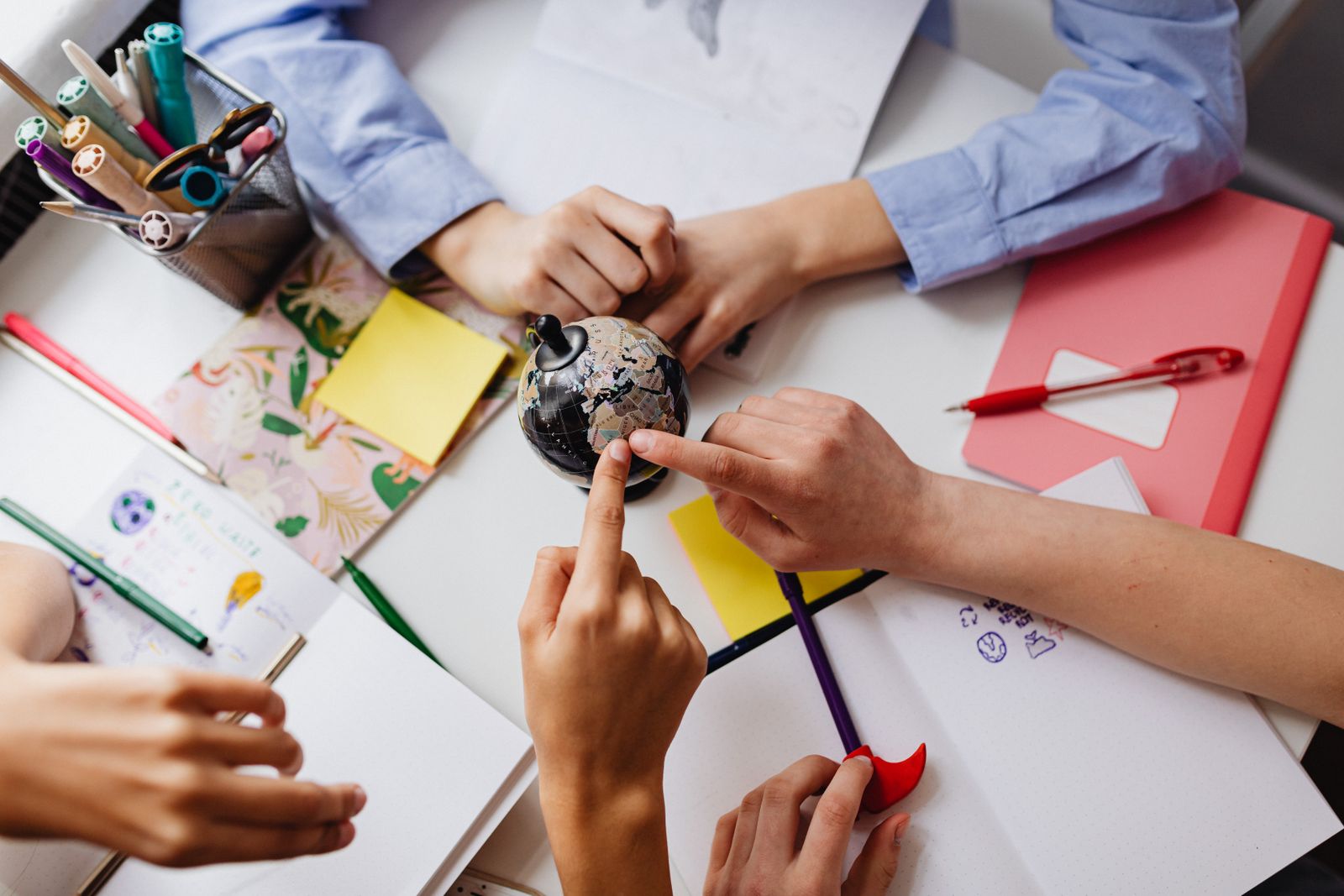 Manos de varios estudiantes sobre un globo terráqueo y cuadernos de trabajo